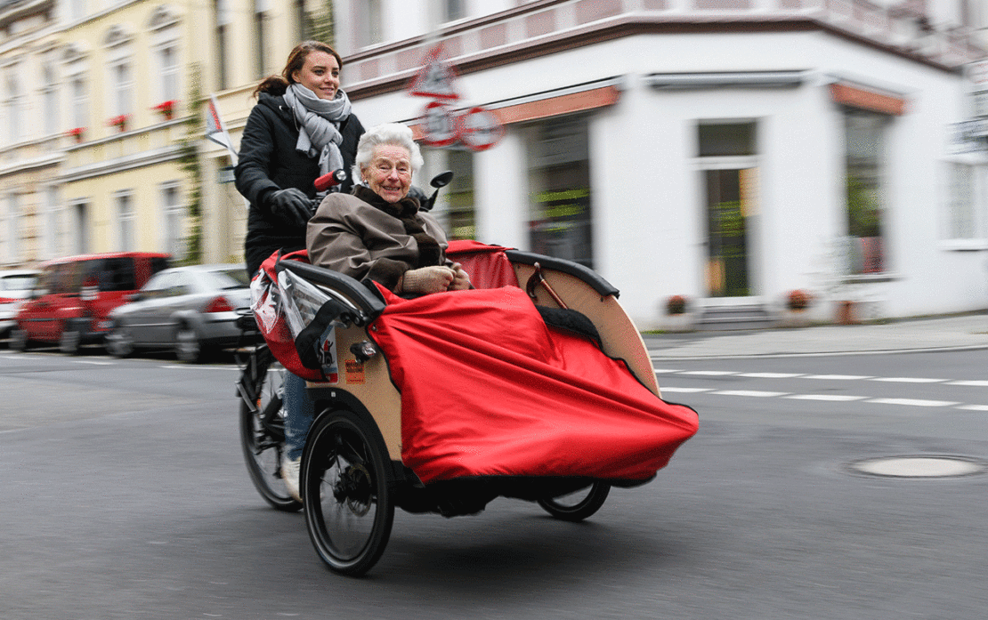 Ehrenamtliche jeden Alters tragen dazu bei, Teilhabemöglichkeiten im lokalen Raum zu schaffen, so auch in Bonn, wo die Ehrenamtliche Caroline Kuhl Seniorin Ruth Nolden in einer Rikscha zum Einkaufen fährt.￼(Foto: KNA)